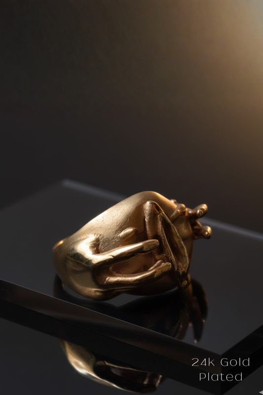 A gold-plated ring with a unique, abstract design sits on a reflective surface against a dark background.