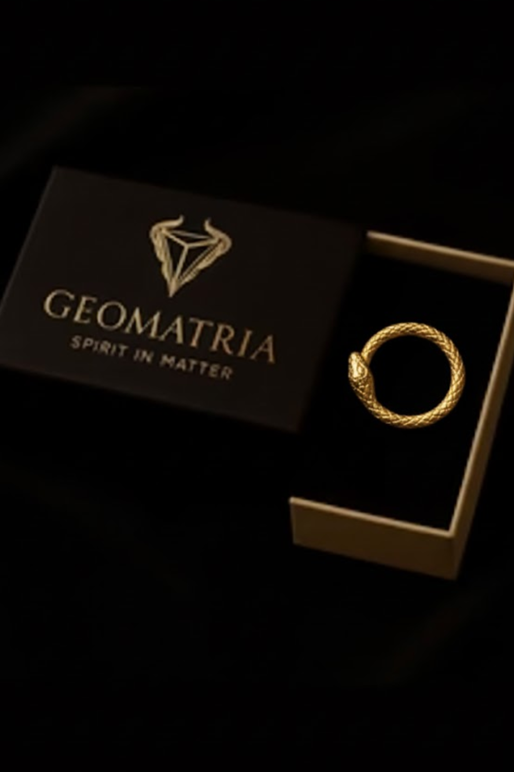 A gold snake ring is displayed in an open gold jewelry box with the text "Geomatria Spirit in Matter" printed on it.