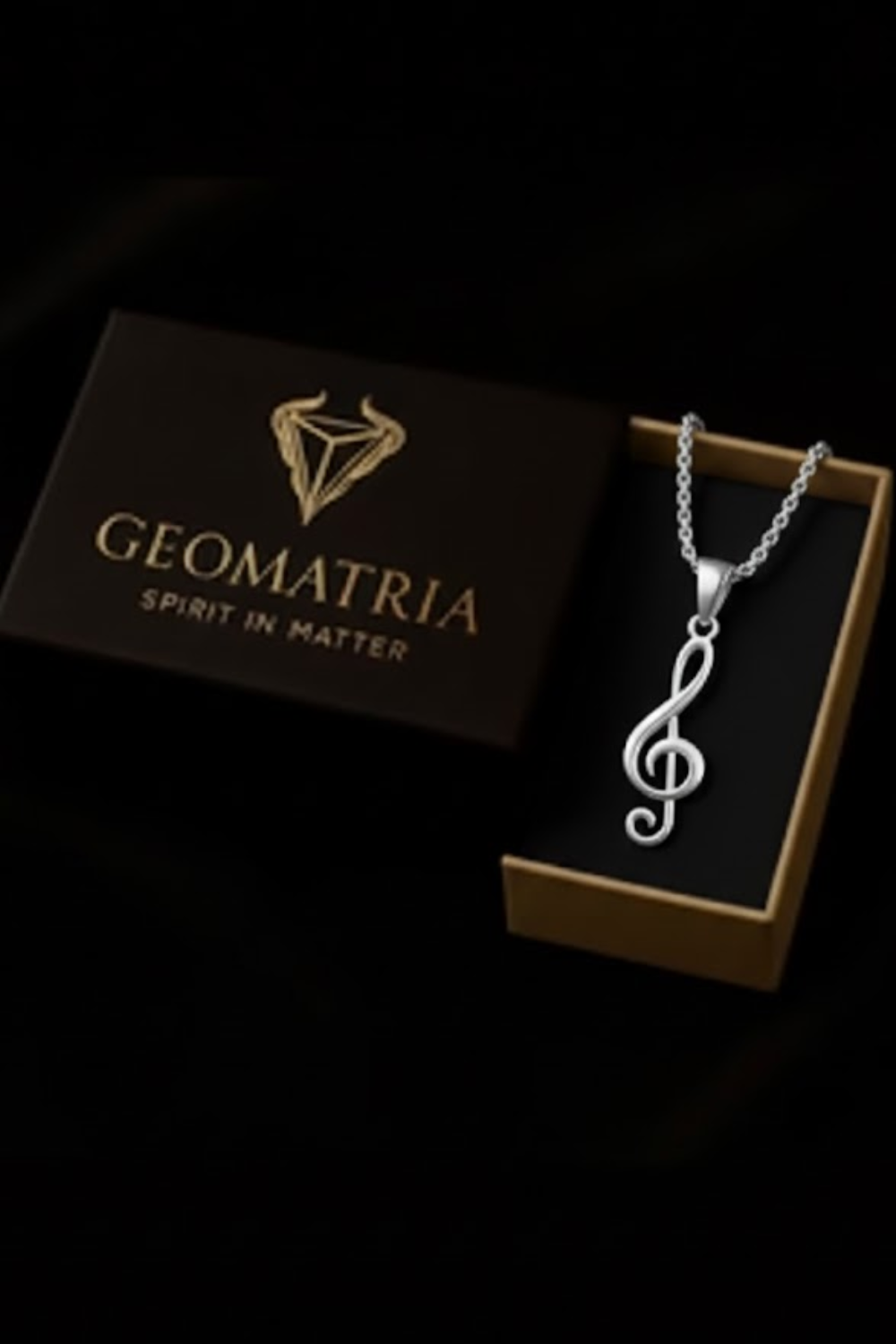A silver necklace with a treble clef pendant is displayed in a gold box with the text "Geomatria" and "SPIRIT IN MATTERS" printed on it.