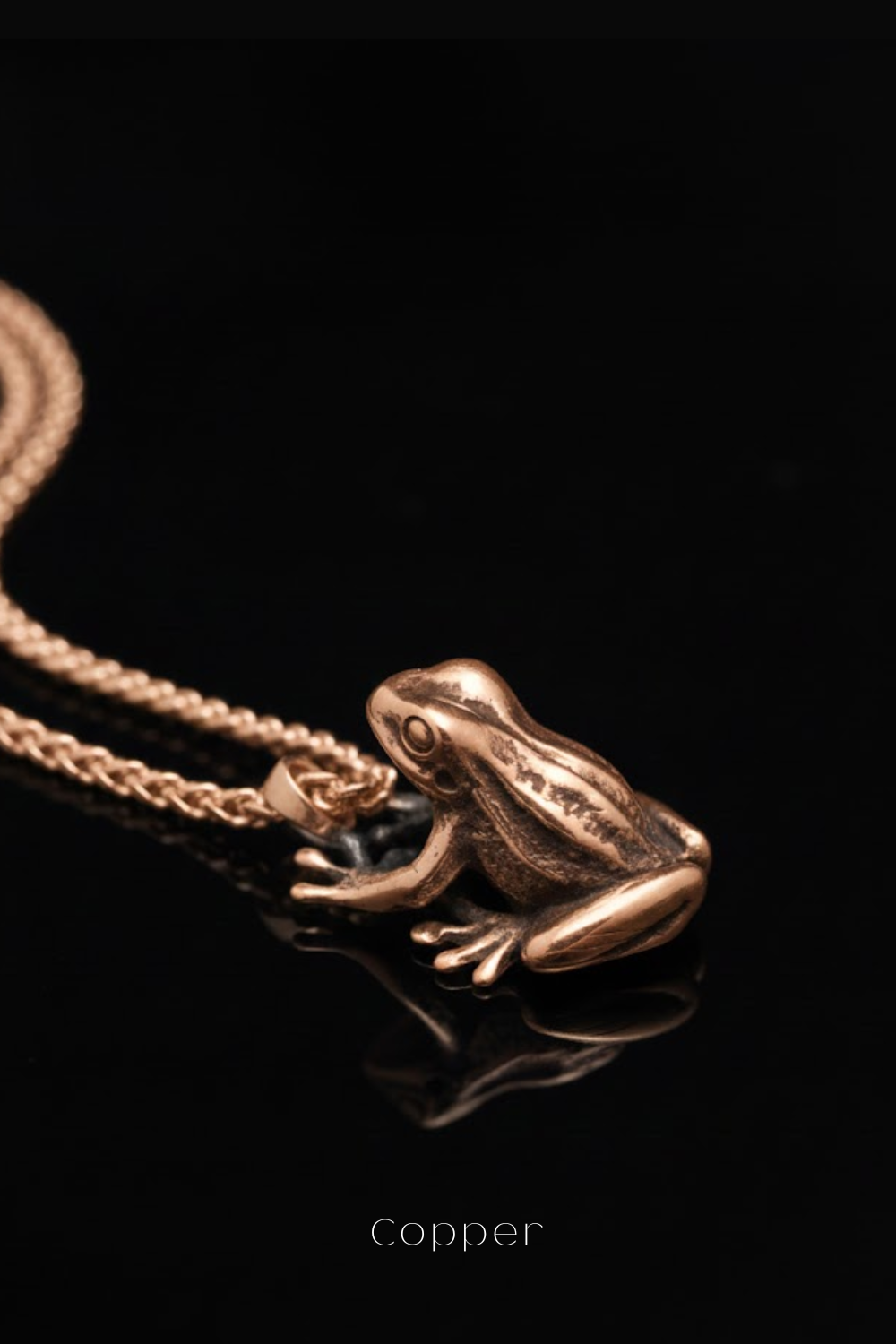 A close-up image of a gold-colored pendant in the shape of a frog, with a chain attached to it, against a black background.