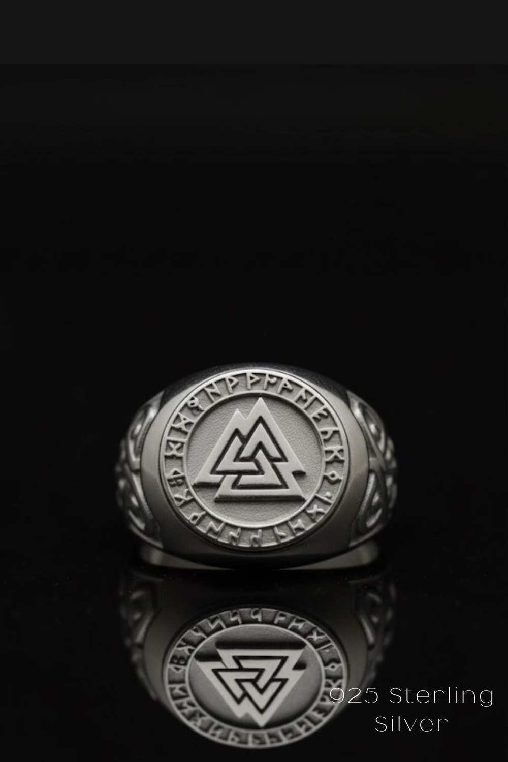 A silver ring with a circular design featuring a triangle and a symbol, placed on a reflective surface against a black background.