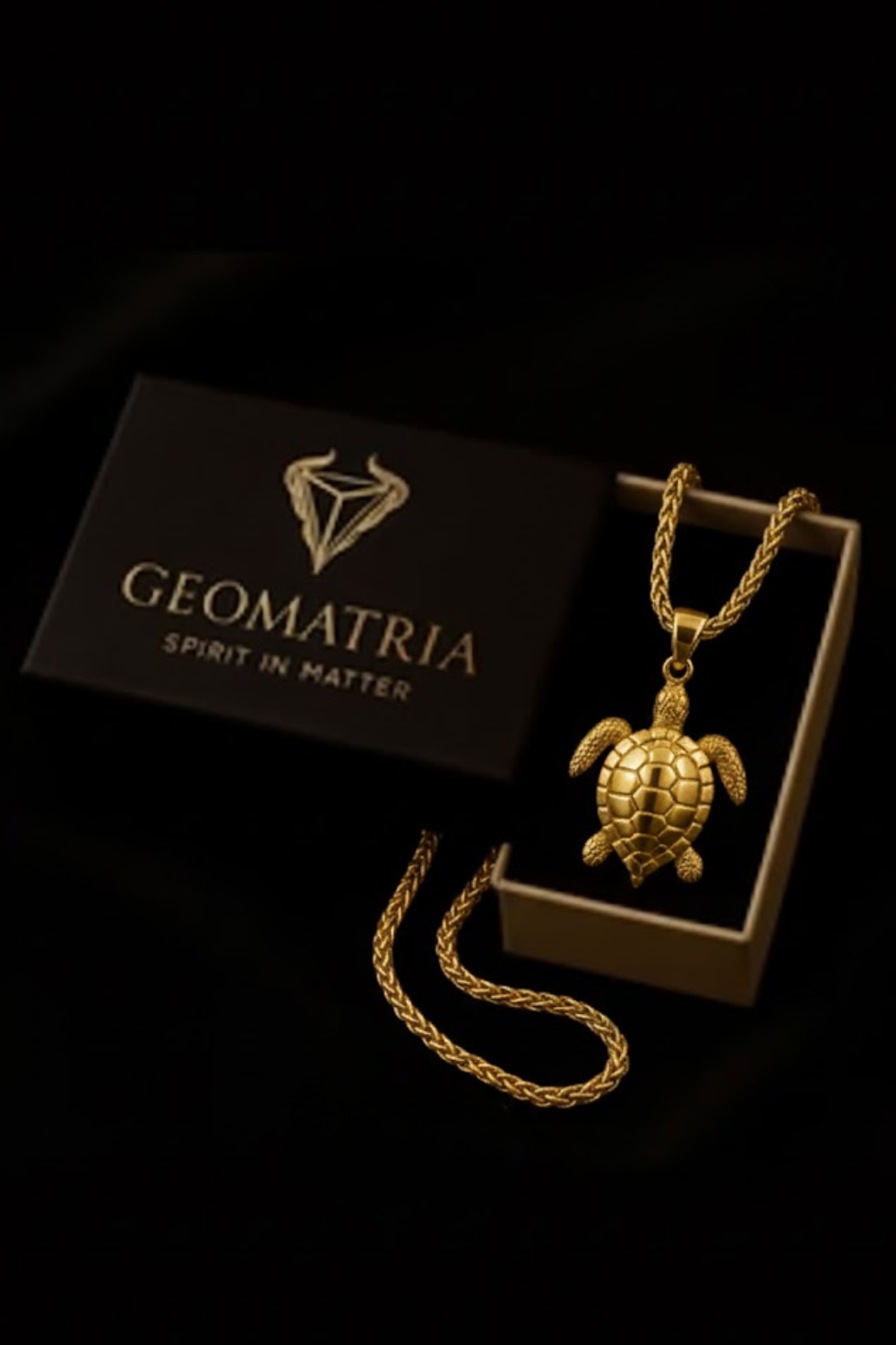 A gold necklace with a pendant in the shape of a turtle is displayed in a black box with the Geomatria logo, accompanied by a gold chain.
