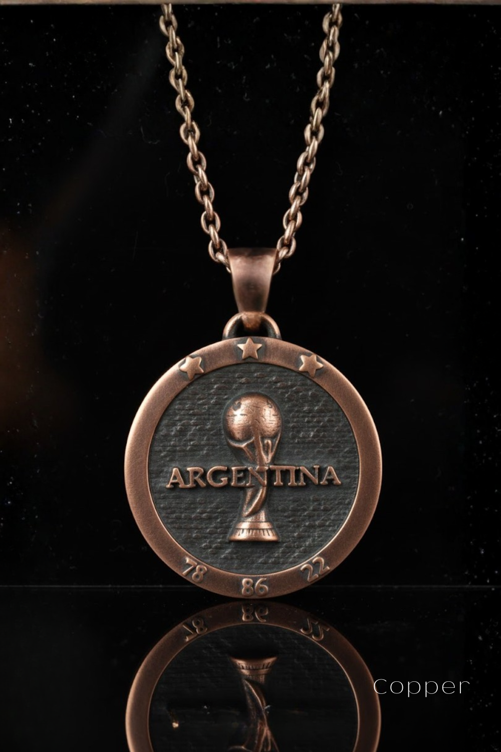 A copper-colored pendant necklace with a circular medallion featuring the word "ARGENTINA" and a soccer ball design, suspended from a chain against a black background.