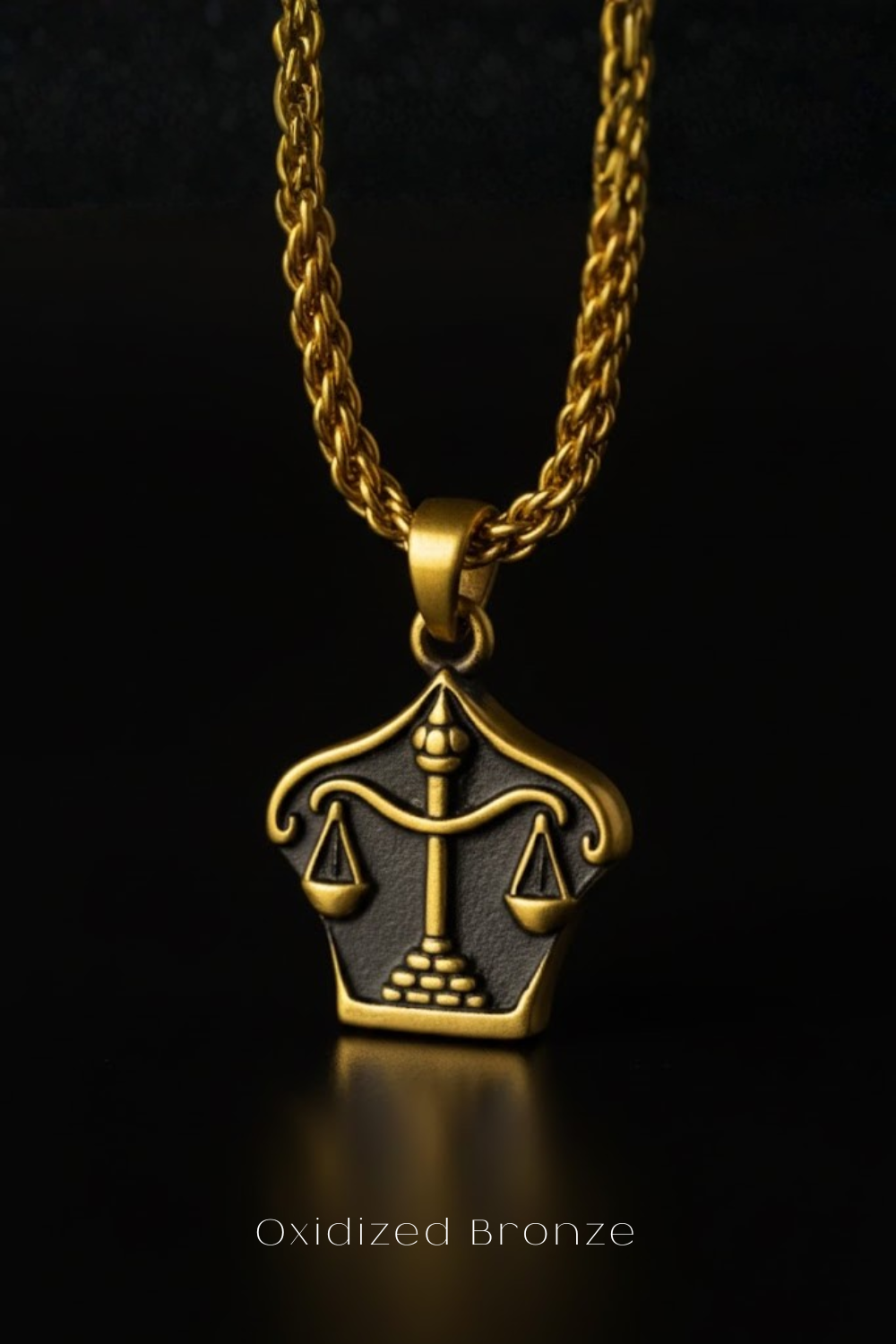 An oxidized bronze pendant with a balanced scale design, suspended from a gold chain against a black background.
