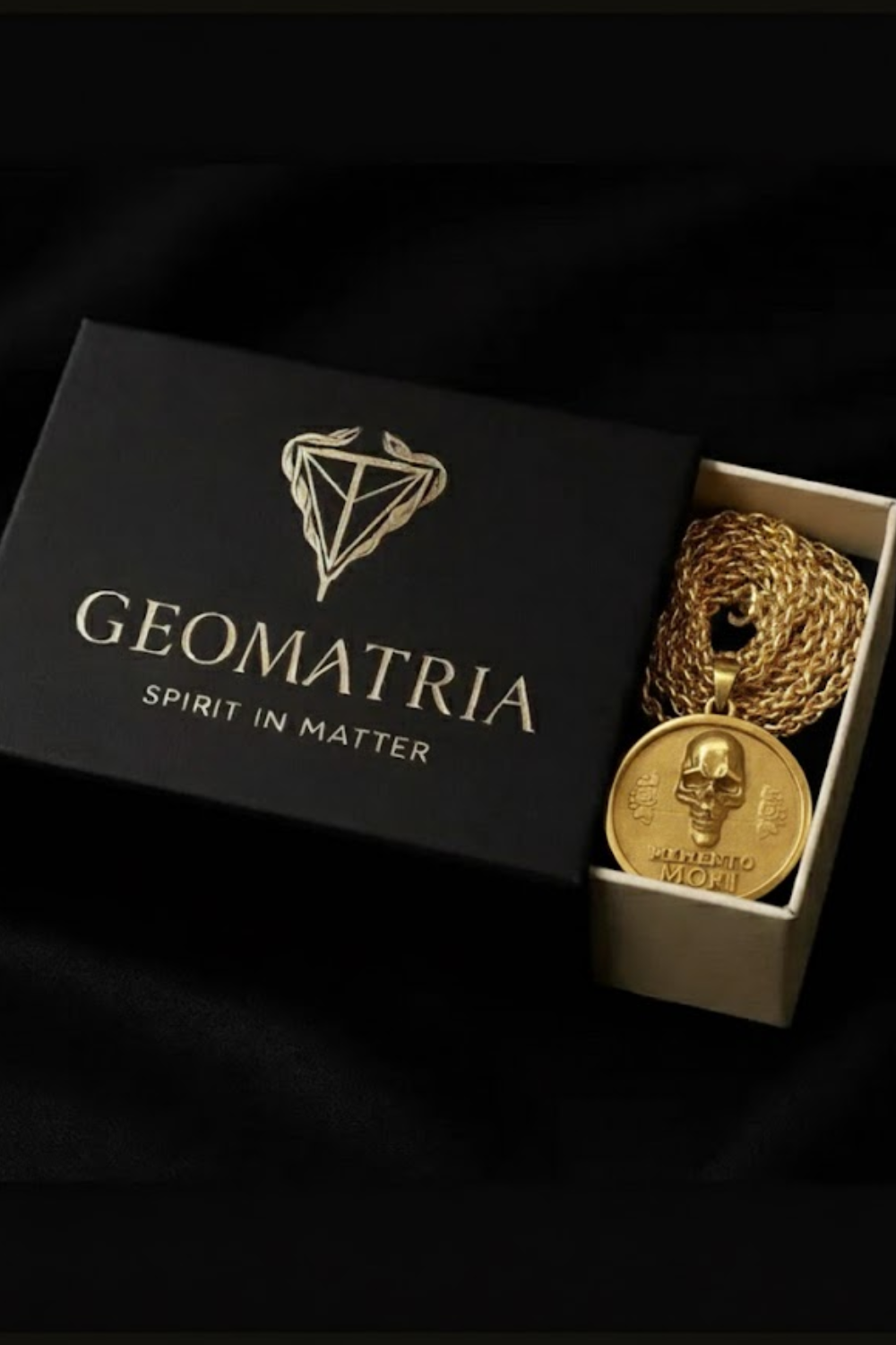 A gold-colored necklace with a pendant featuring a skull design, placed inside a black box with the word "Geomatria" printed on it.