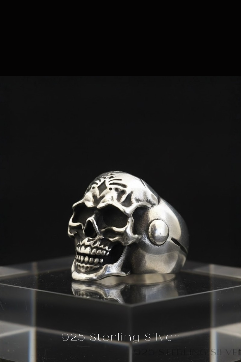 A silver skull ring is displayed on a clear glass stand against a black background.