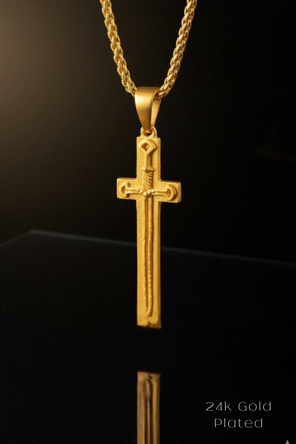 A 24k gold plated cross pendant is displayed against a black background, reflecting on a surface below.