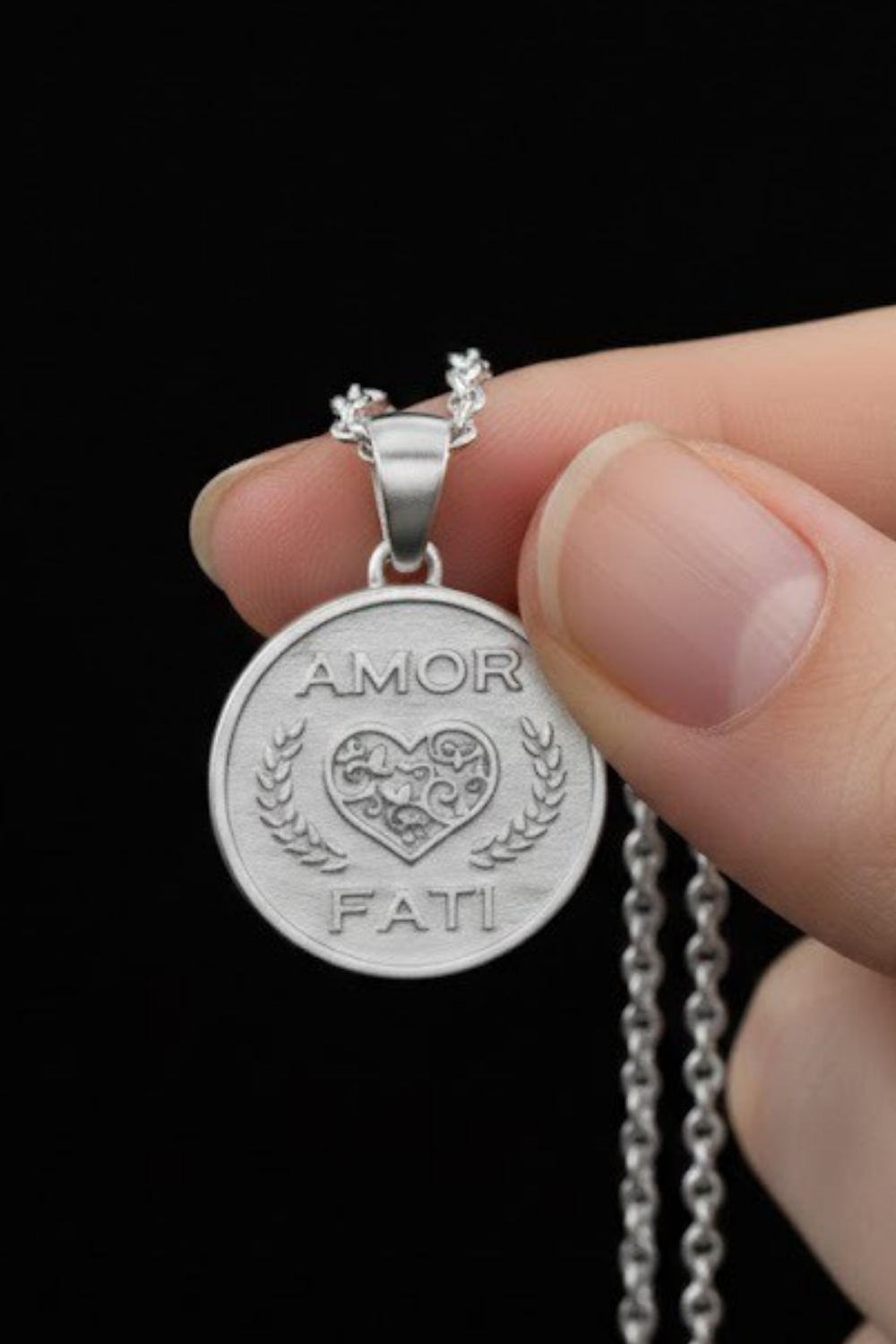A hand holding a silver pendant with the words "AMOR FAMI" engraved on it.