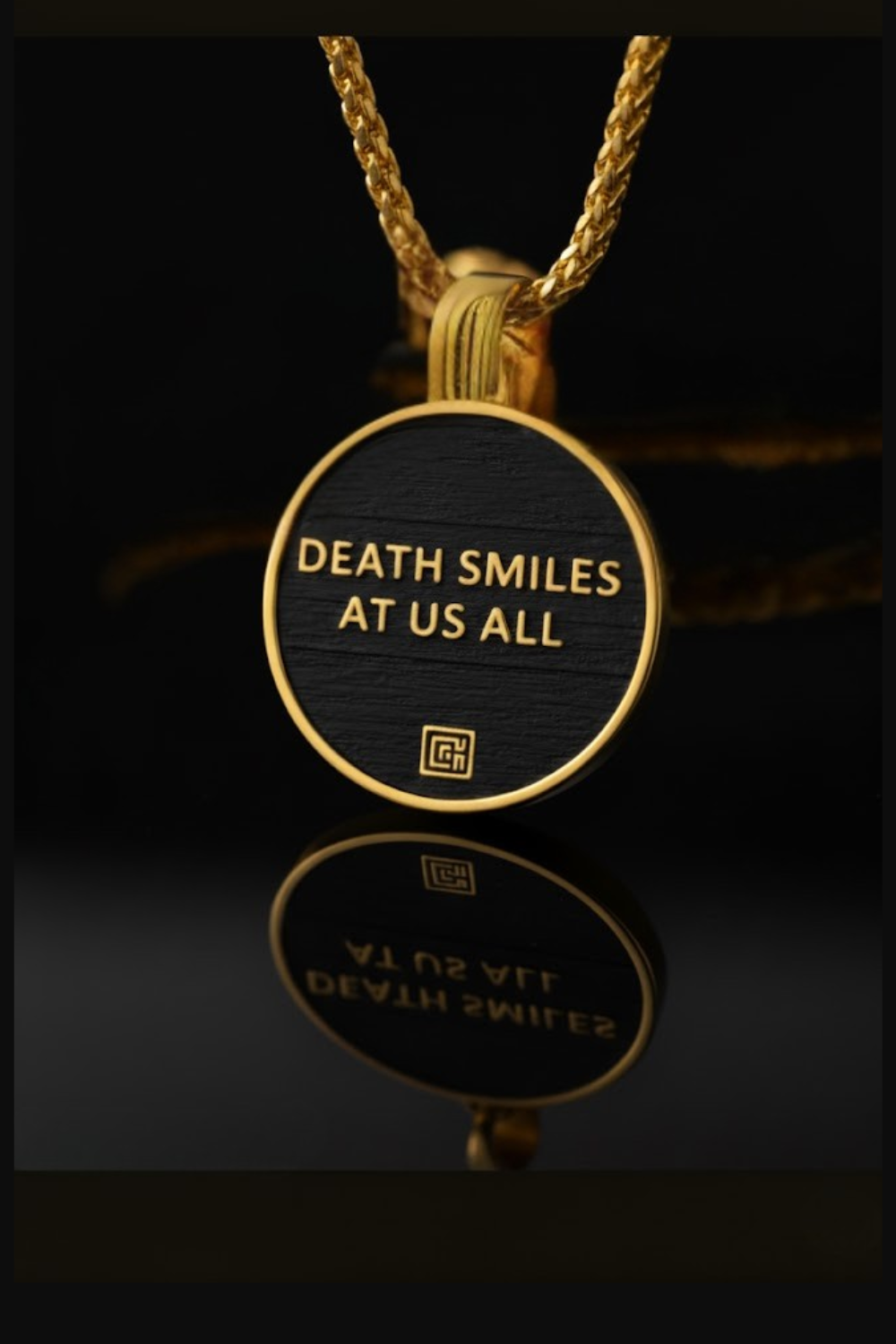 A gold-colored pendant with a black circular background and the text "DEATH SMILES AT US ALL" printed on it.