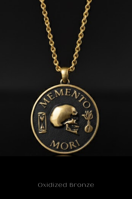 a gold-colored pendant necklace with a circular design on a black background. The pendant has the Latin phrase "Memento mori" inscribed on it, which translates to "Remembering the dead" in English.