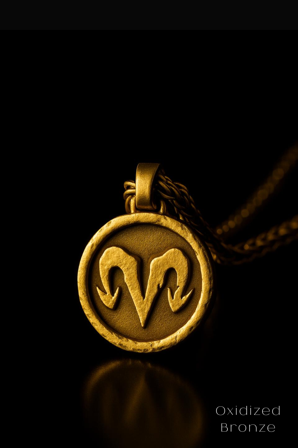 A close-up image of a gold-colored pendant with a circular design featuring a stylized symbol, such as a pair of horns or a stylized letter, against a black background.