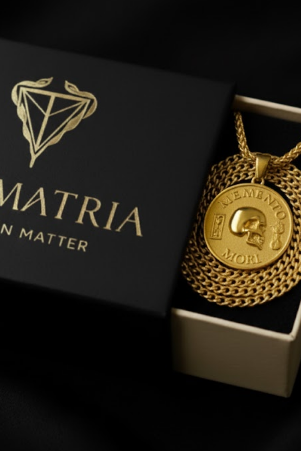 a gold-colored pendant necklace with a skull pendant, placed inside a black box with the word "MATRIIA" printed on it.
