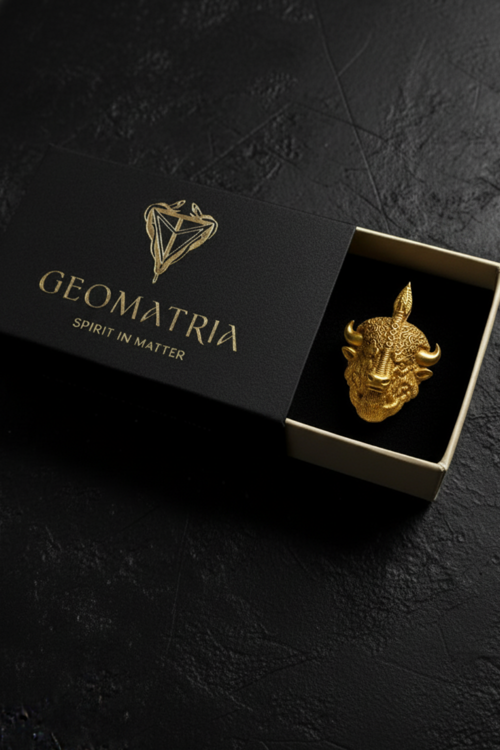 A gold bull head brooch is displayed in a black box with the text "Geomatria" and "SPIRIT IN MATTER" printed on it.