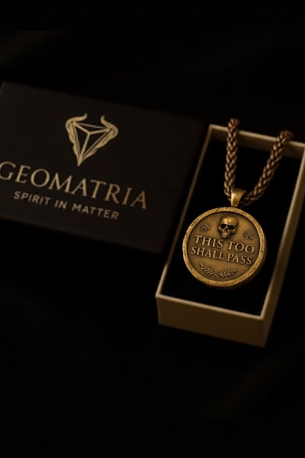 a gold-colored pendant necklace with a skull pendant, displayed in a black box with the text "Geomatria Spirit in Matter" printed on it.