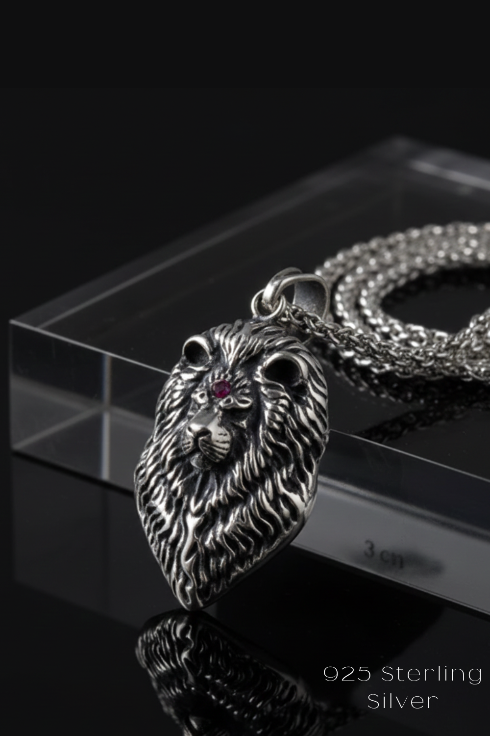A silver pendant in the shape of a lion's head with a red gemstone, displayed on a clear glass stand.