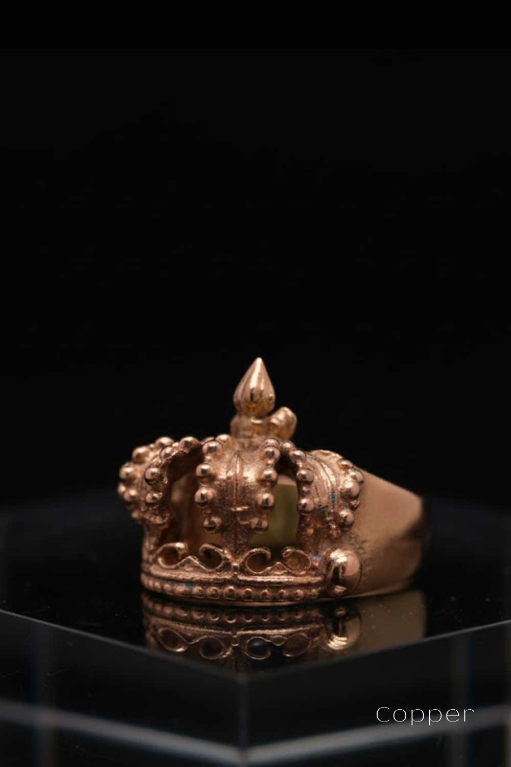 A gold-colored ring with a decorative crown-like design sits on a reflective surface against a black background.