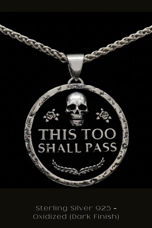 A silver pendant necklace with a circular design featuring a skull and the text "THIS TOO SHALL PASS" in a bold, black font.