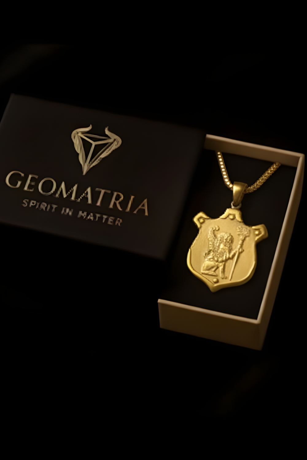 A gold-colored pendant with a shield-shaped design is displayed in a black box with the text "Geomatria" and "SPIRIT IN MATER" printed on it.