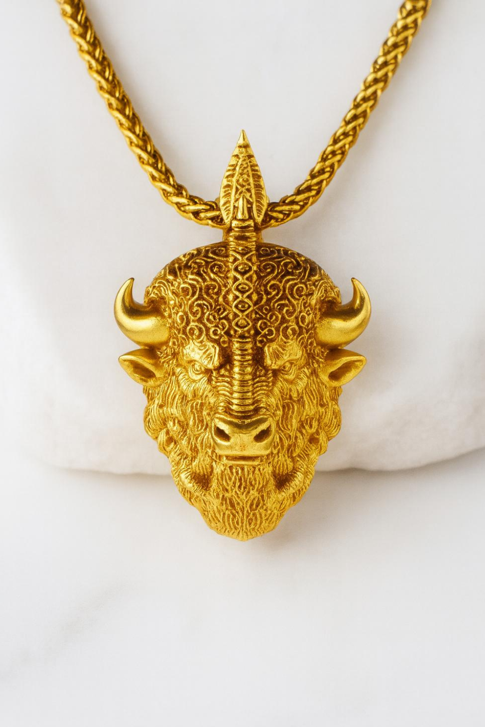A gold-colored pendant in the shape of a buffalo head with a pointed top and curved horns.