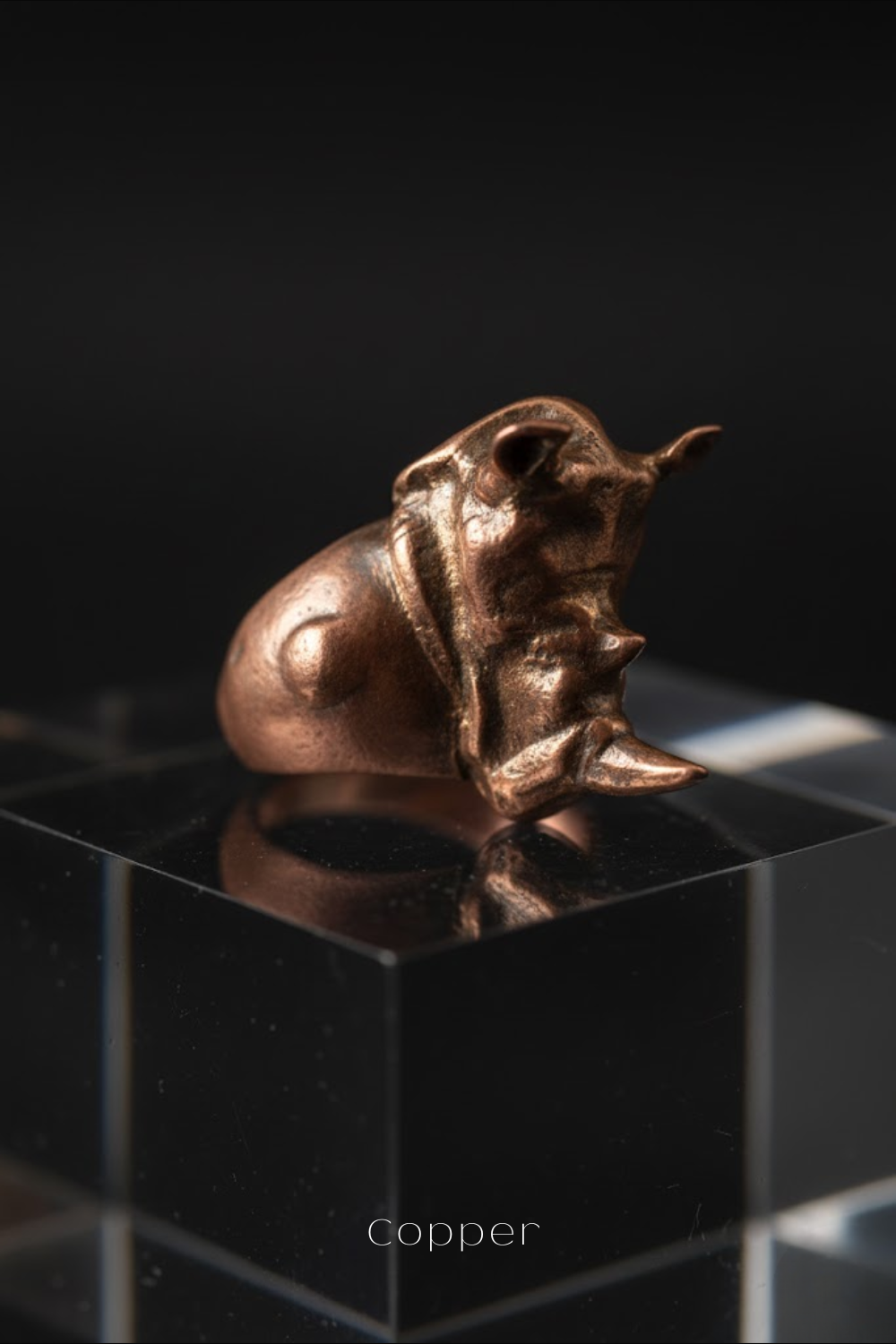 A copper-colored metal sculpture of a rhinoceros is displayed on a clear glass stand against a black background.