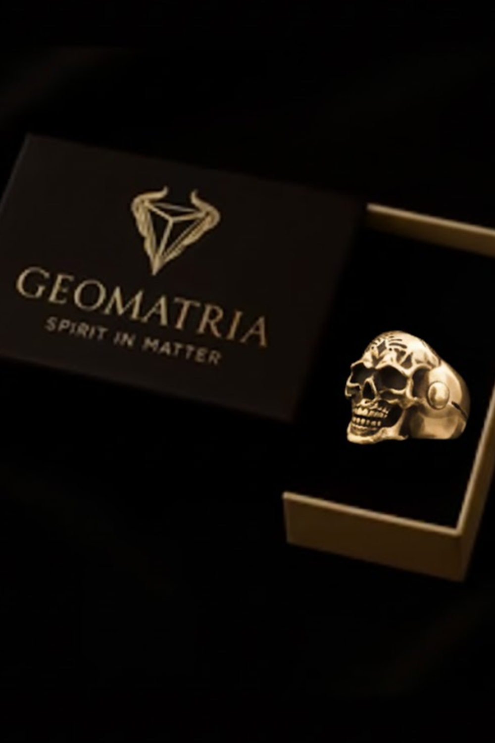 A gold-colored skull ring is displayed in a black box with the text "Geomatria Spirit in Matter" printed on it.