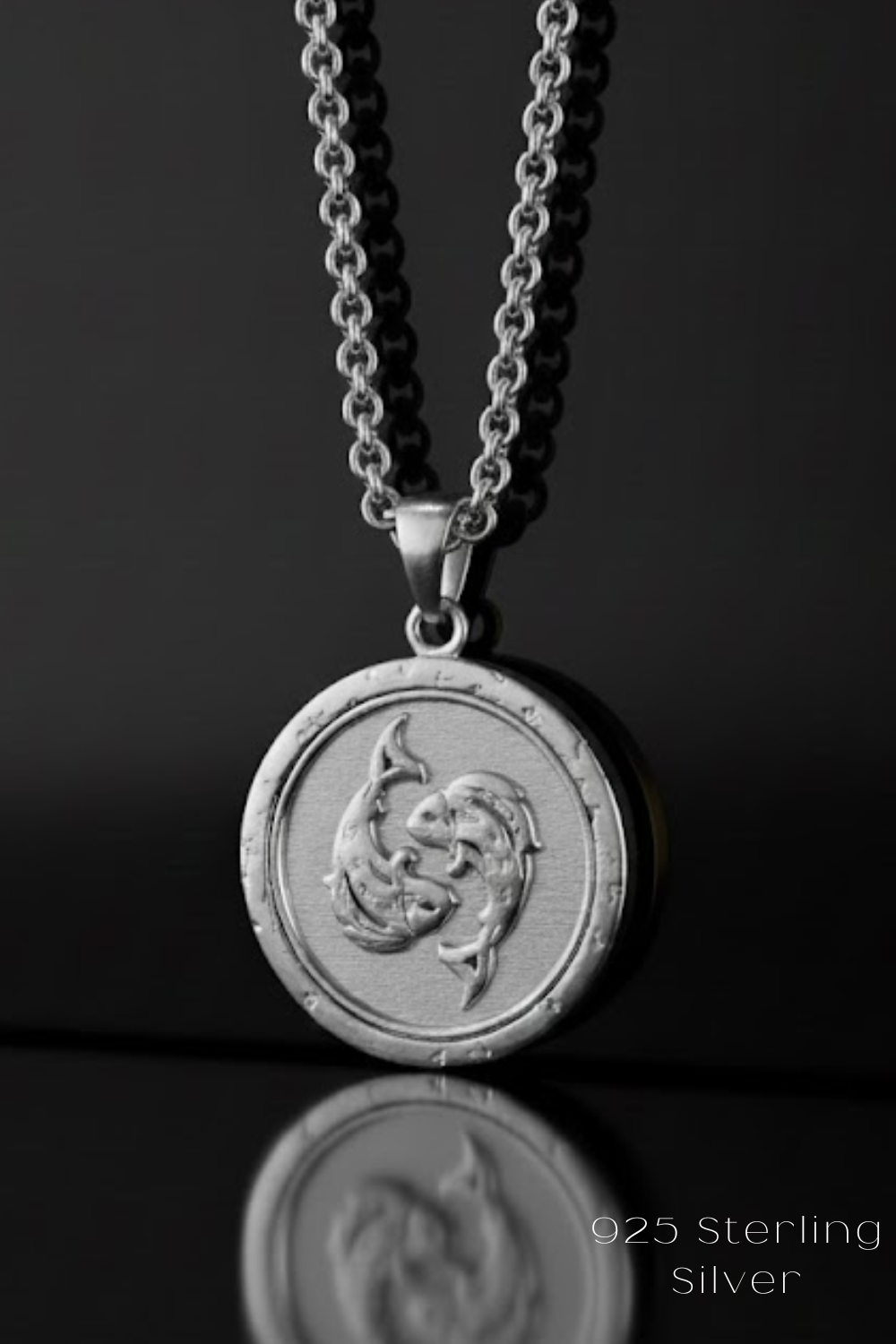 A silver-colored pendant necklace with a circular medallion featuring a fish design, suspended from a chain against a dark background.