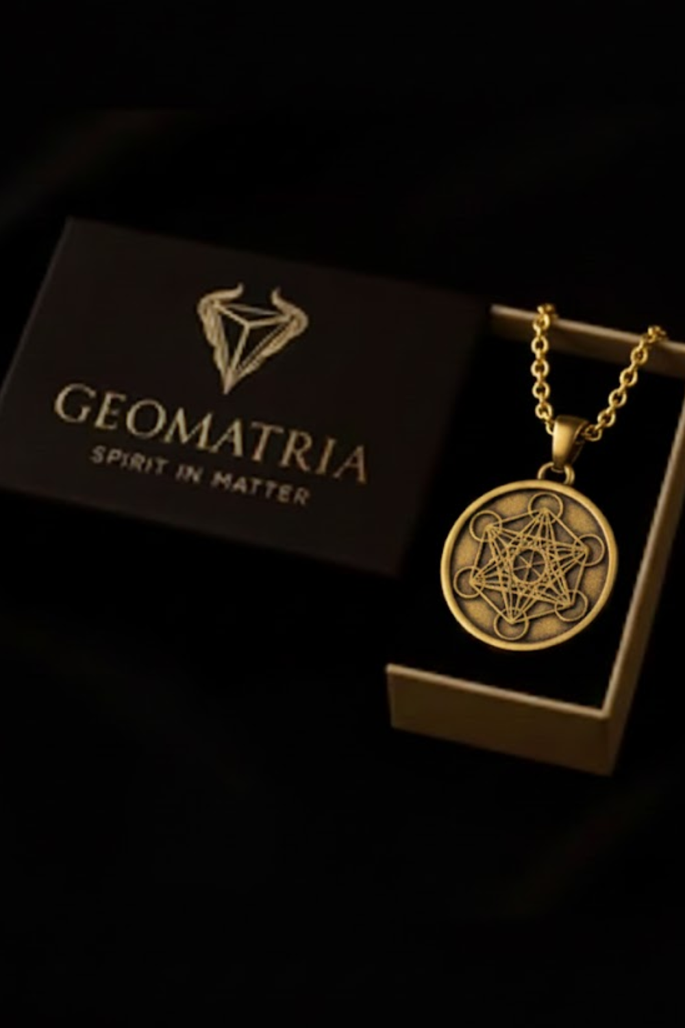 A gold-colored necklace with a pendant featuring a geometric design, placed in a black box with the text "Geomatria" printed on it.