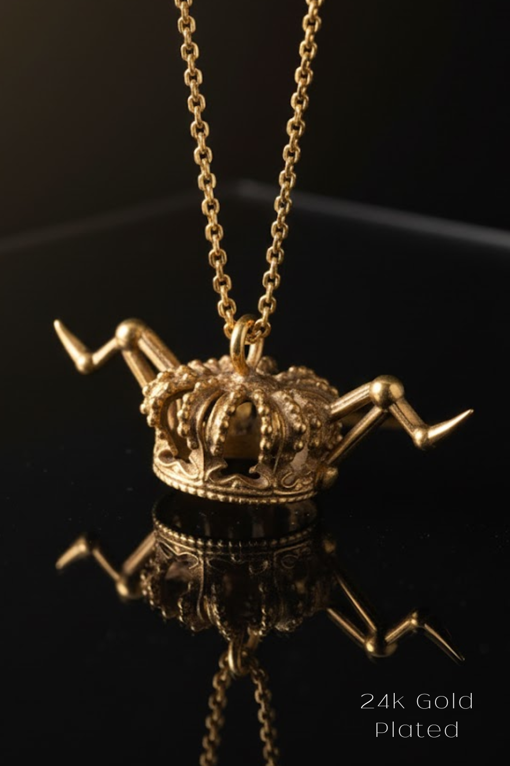 A gold-plated necklace with a crown-shaped pendant is displayed against a black background, reflecting on the surface below.