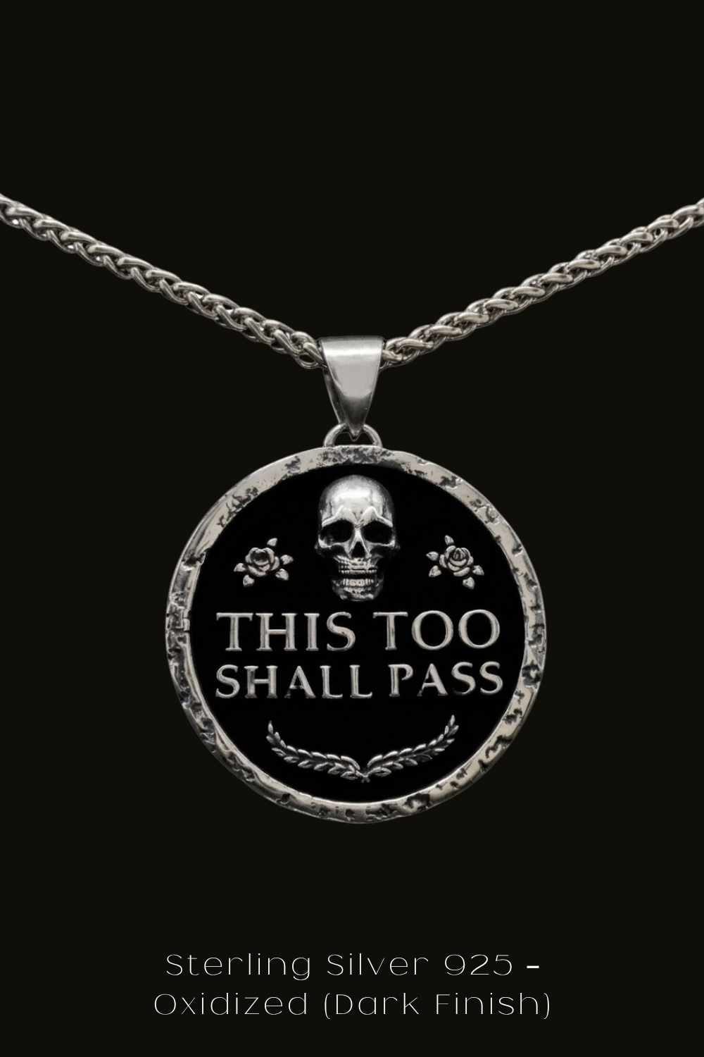 A silver pendant necklace with a circular design featuring a skull and the text "THIS TOO SHALL PASS" in a bold, black font.