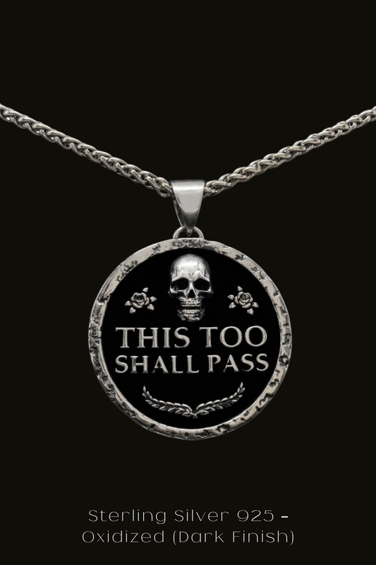 A silver pendant necklace with a circular design featuring a skull and the text "THIS TOO SHALL PASS" in a bold, black font.