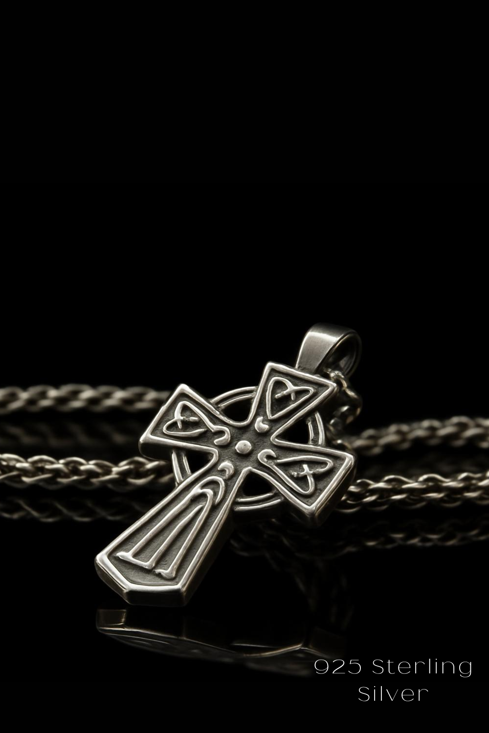 A silver cross pendant with intricate designs is displayed against a black background, accompanied by a silver chain.