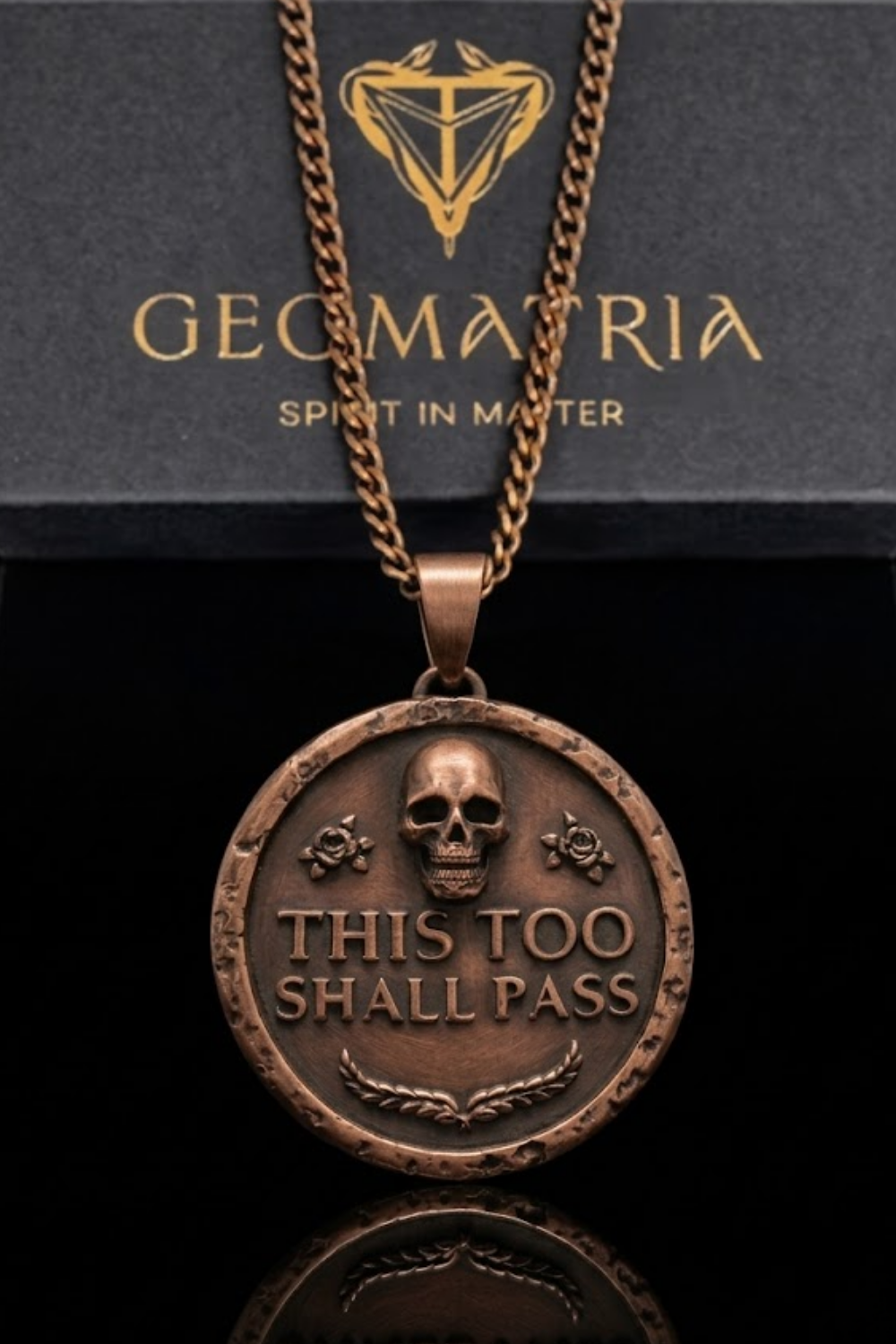 A large, round, metal pendant with a skull design and the text "This Too Shall Pass" engraved on it. The pendant is hanging from a chain.